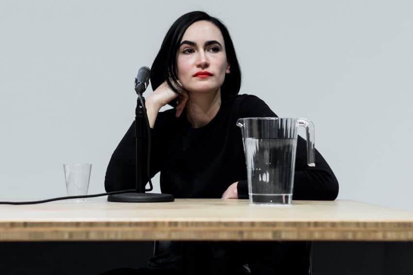 Architect Frida Escobedo sitting at a butcher block table with a tabletop microphone stand, a filled glass pitcher of water. She is sitting with her right hand crooked against the right side of her face and staring off into the distance.