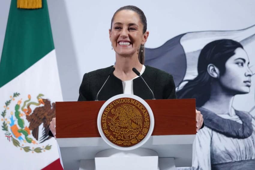 President Claudia Sheinbaum standing at the presidential podium at the National Palace with a wide smile as she gazes out at reporters off camera. Behind her is a large Mexican flag on a post and a wall with the logo of Sheinbaum