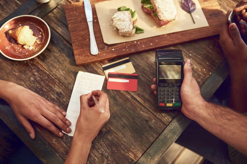 A server holds a payment terminal next to a restaurant table, while a diner signs their bill