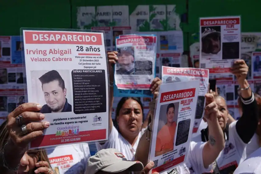 Mexican men and women at a protest in Mexico City, holding up laminated missing persons posters of their loved ones. All the posters say at the top