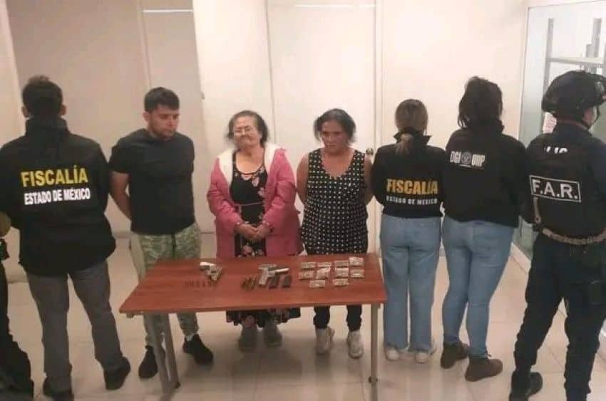 An elderly woman in a pink coat stands behind a table with a gun and packets of powder, next to two younger handcuffed people and several police agents