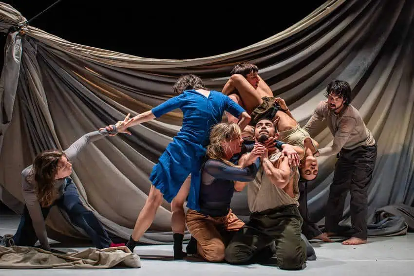 A stage in which six actors are in a performance, some kneeling, some standing, but all holding each other in some way. The man in the center of the group is looking upward as an actress holds up his chin