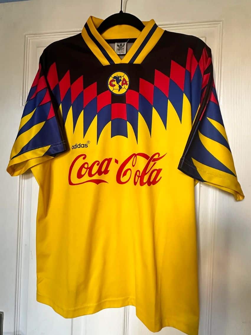 Una camiseta de uniforme del equipo de fútbol amarillo para el equipo de fútbol Club America en la Ciudad de México, en una percha que cuelga de una puerta blanca interior en una casa residencial. Tiene diamantes azul marino en un patrón en zigzag, el logotipo de Club America, justo debajo del cuello en V, y los nombres de Coca Cola y Adidas marcados hacia el Centro de Jersey