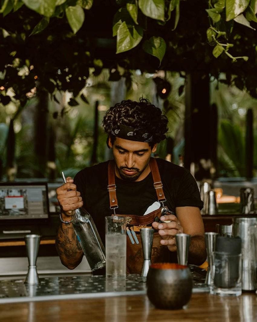 Un camarero mexicano que usa un delantal de cuero sobre una camiseta negra y un pañuelo que se sufre el cabello está en el medio de mezclar un cóctel de aspecto claro en un vaso alto.