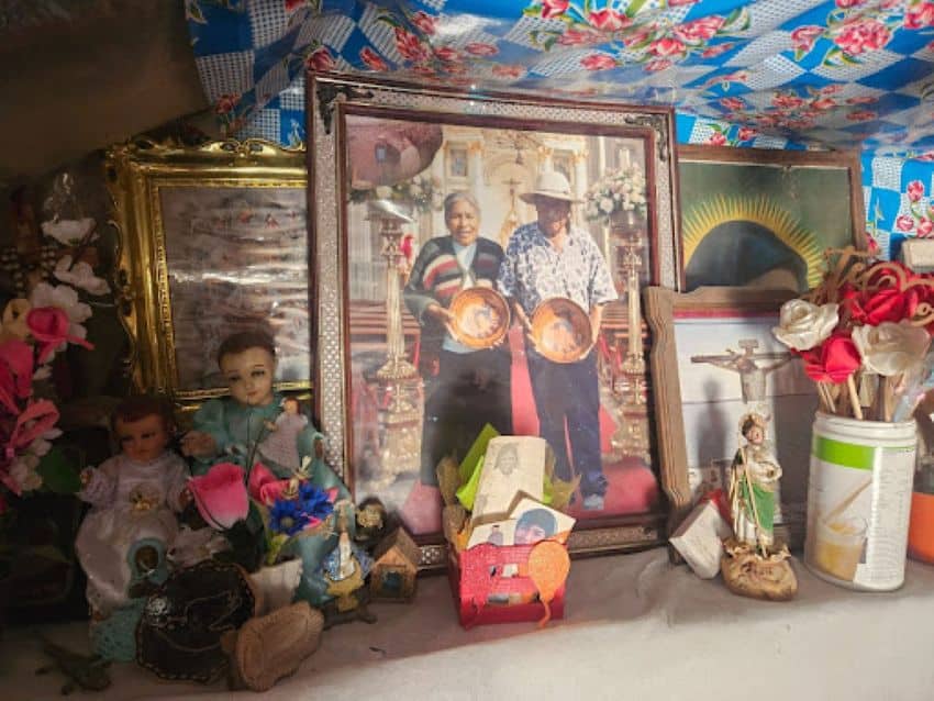 Una mesa con algunas fotos y pinturas enmarcadas diferentes, algunas de la naturaleza cristiana. En el centro hay una foto de un hombre mayor y una mujer que sostiene tazones de cerámica artesanal frente a ellos.