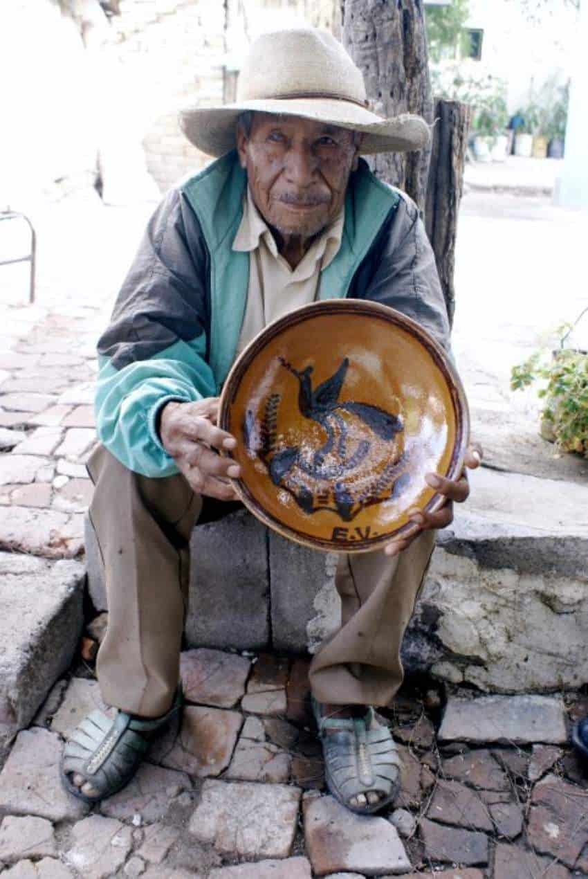 Un anciano que lleva un sombrero de paja se sienta en un muro de piedra baja en una calle de la ciudad y sostiene ante él un tazón de cerámica que hizo a mano.