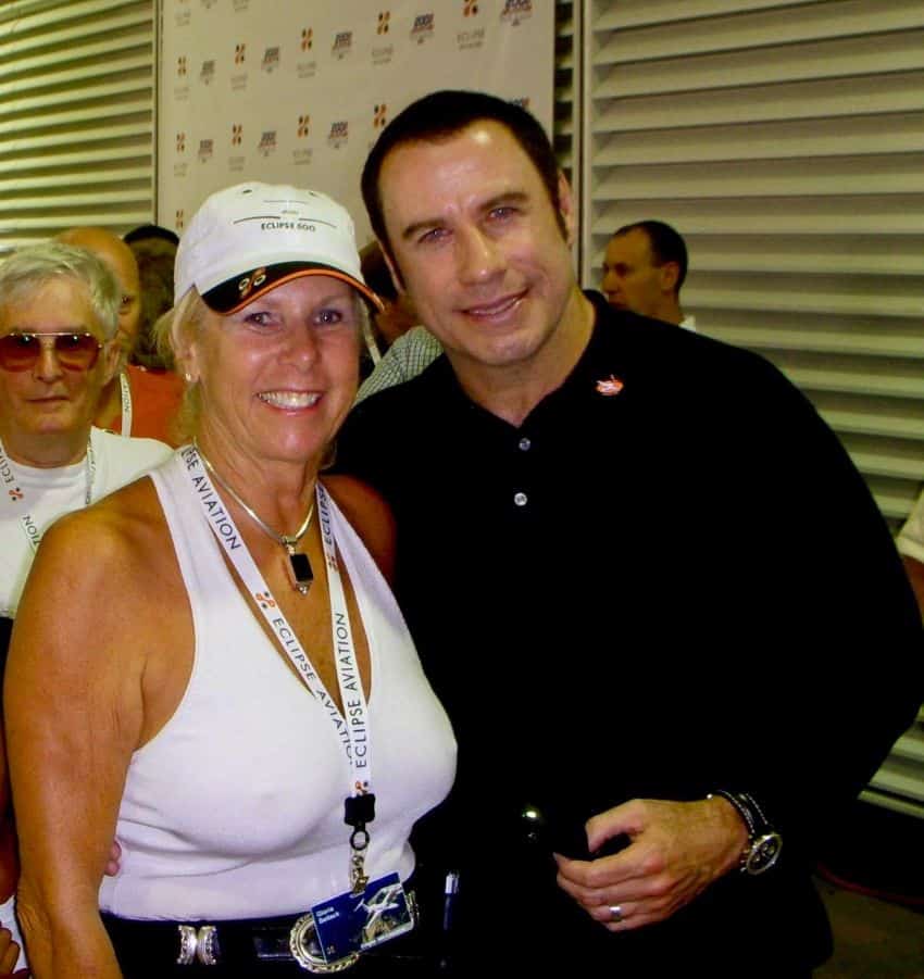 Una mujer rubia de mediana edad posa con el actor John Travolta para una selfie en un evento de aviación Eclipse en la década de 1980. Otros invitados miran detrás de ellos.