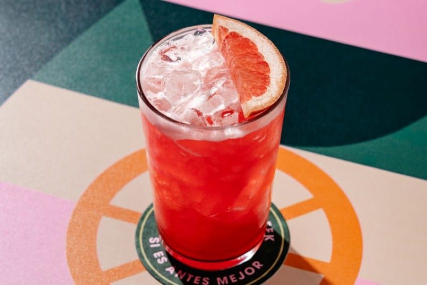Alto vaso alto con una bebida de color de toronja. Hay una cuña de toronja que descansa sobre los cubitos de hielo de la bebida flotando en la parte superior del vaso.