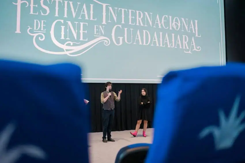 Un hombre y una mujer se encuentran en un escenario en un teatro bajo una gran pantalla de proyección que tiene el logotipo para el Festival Internacional de Cine de Guadalajara en español.