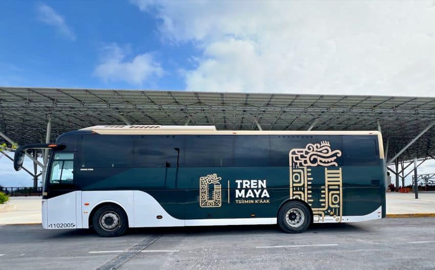 Autobús de pasajeros con librea de tren maya frente a una estación de tren, durante el día
