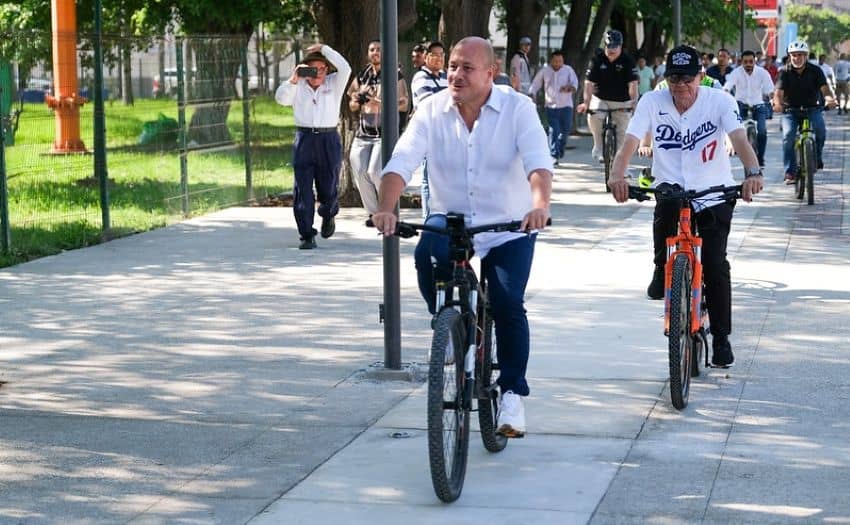 El gobernador de Jalisco, Enrique Alfaro, montando una bicicleta en un nuevo carril para bicicletas en Puerto Vallarta. Está a la cabeza con una camisa de algodón blanca con un botón y pantalones negros y zapatillas blancas en una bicicleta de montaña negra. Detrás de él a distancias variables, hay otros ciclistas. En el fondo, un hombre con una camisa de vestir y pantalones negros lo lleva imágenes con una cámara.