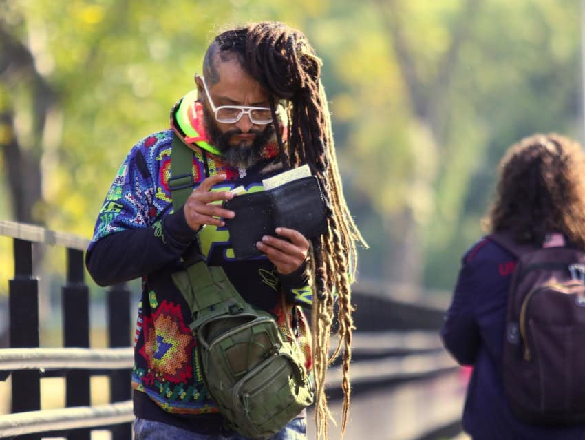 Un joven mexicano con un suéter colorido y gafas de lectura blanca y que luce rastas de la cintura para su cintura sobre el contenido de su billetera en el campus fuera del centro de campus de la Universidad Nacional Autónoma de México. Cerca, una joven estudiante que lleva una chaqueta y una mochila camina en la otra dirección