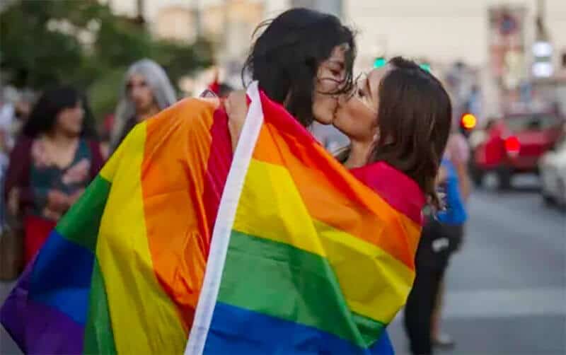 Dos mujeres besándose en la calle envueltas en una bandera multicolor de orgullo gay de arco iris.