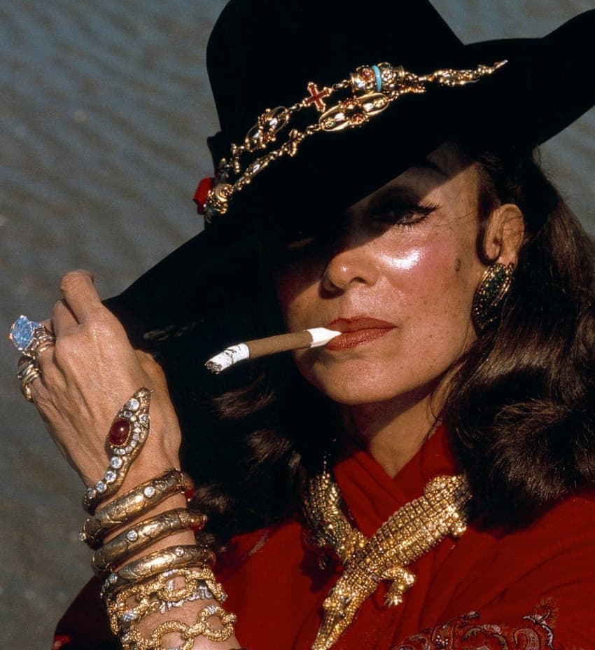María Félix fumando mientras usa un gran sombrero negro y libras de joyas de oro y piedras preciosas en forma de reptiles