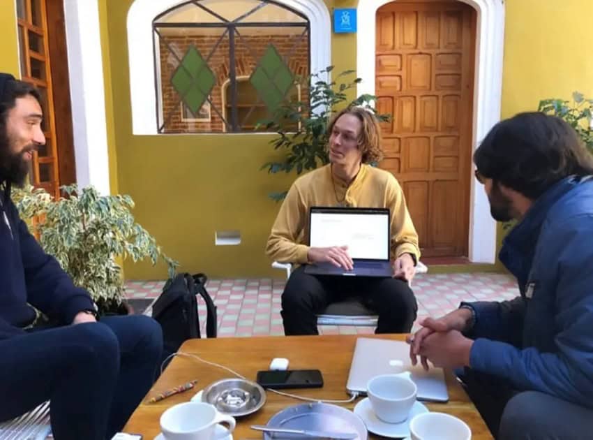 Tres jóvenes sentados en una mesa de café en un patio mexicano. Uno tiene una computadora portátil abierta en su regazo y otra tiene una computadora portátil en la mesa junto a su taza de café. El tercero es escuchar al hombre con la computadora portátil en su regazo.