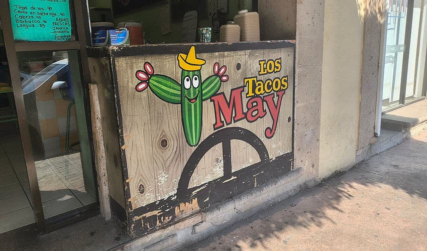 Un letrero de taquería que presenta un cactus de dibujos animados de saguaro con ojos y una sonrisa y un sombrero Paisano de estilo mexicano.
