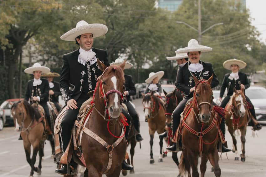 Hombres mexicanos de diferentes edades a caballo con elegantes trajes de charro, montando en procesión a través de las calles de Guadalajara a medida que los automóviles en el fondo ceden a ellos.