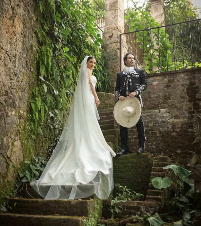 Una pareja de bodas, la novia con un vestido de novia blanco completo y el hombre con una regalía de charro mexicana, posando en algunos escalones de piedra por un muro de piedra cubierto de hiedra y musgo.
