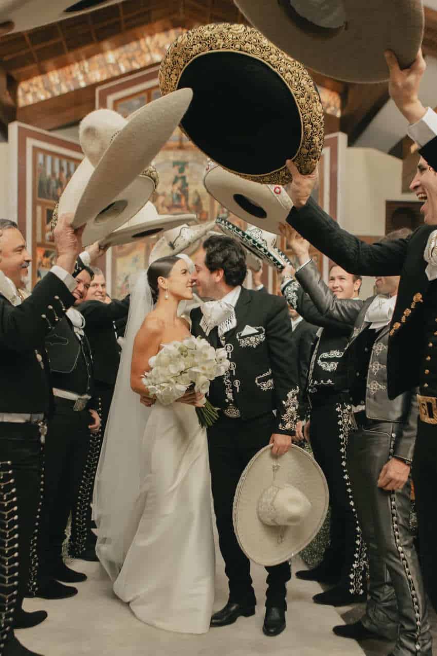 Un novio besándose en el pasillo de una iglesia bajo una fila de sombreros mexicanos sostenidos por dos líneas opuestas de hombres mexicanos con trajes de charro. El novio también lleva un traje de charro. Los Charros a cada lado de la pareja están sonriendo y riendo.