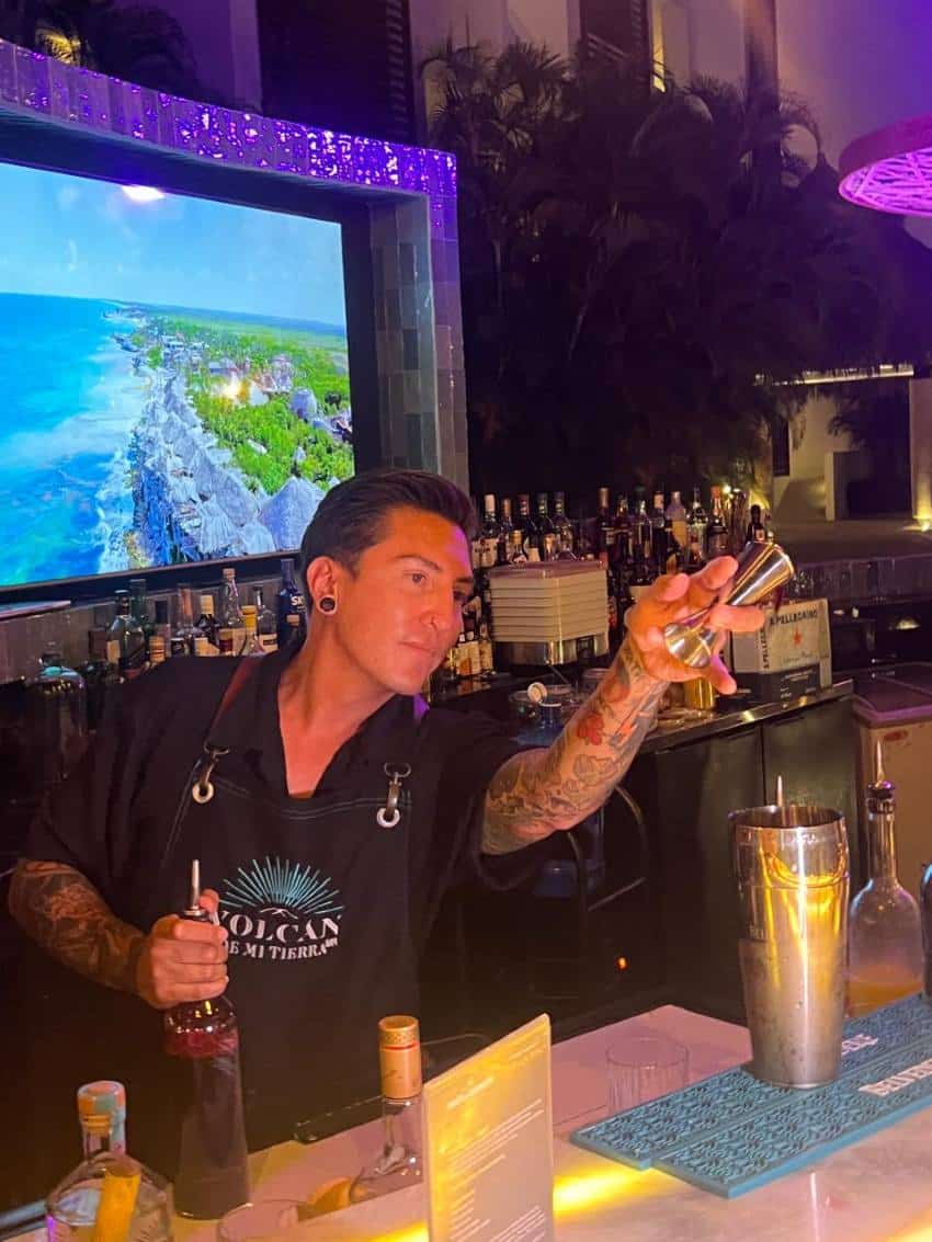 Bartender en una barra sosteniendo una botella de licor con una boquilla de vertido sobre él y sosteniendo en la otra mano una medida de disparo. Está haciendo un gesto con la medida de disparo a alguien o algo fuera de la cámara como si recibiera un pedido de un patrón.