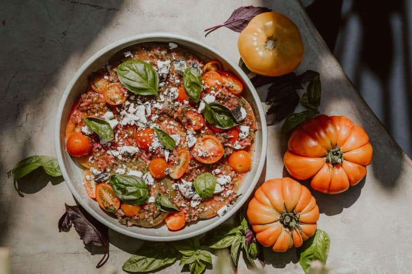 Tomates asados ​​exhibidos ingeniosamente en un plato para servir con hojas de albahaca fresca, rodeados de tomates de reliquia gordos.