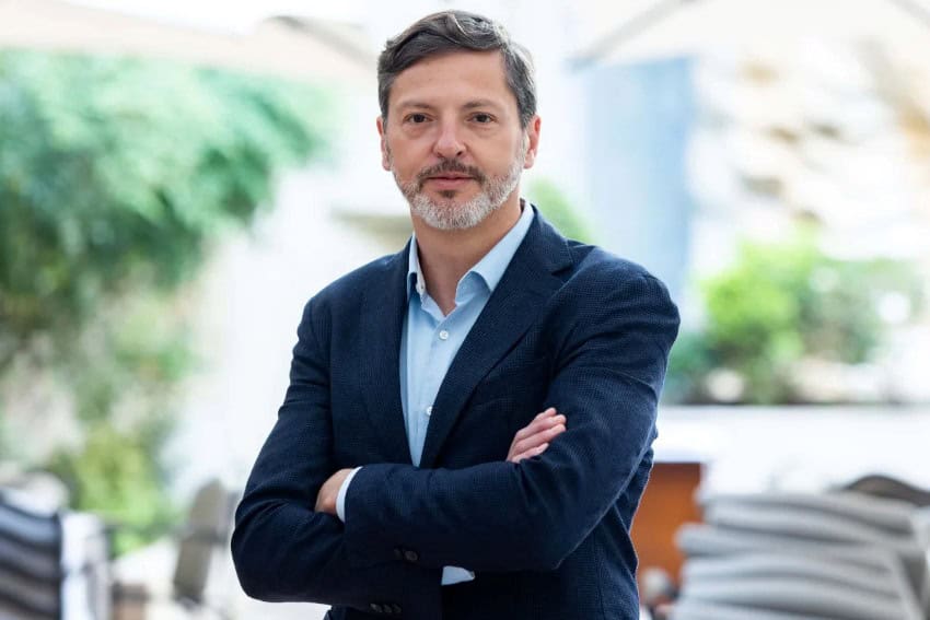 Un hombre mexicano con una blazer azul y una camisa de vestir azul claro se encuentra frente a la cámara con los brazos doblados sobre su cuerpo.