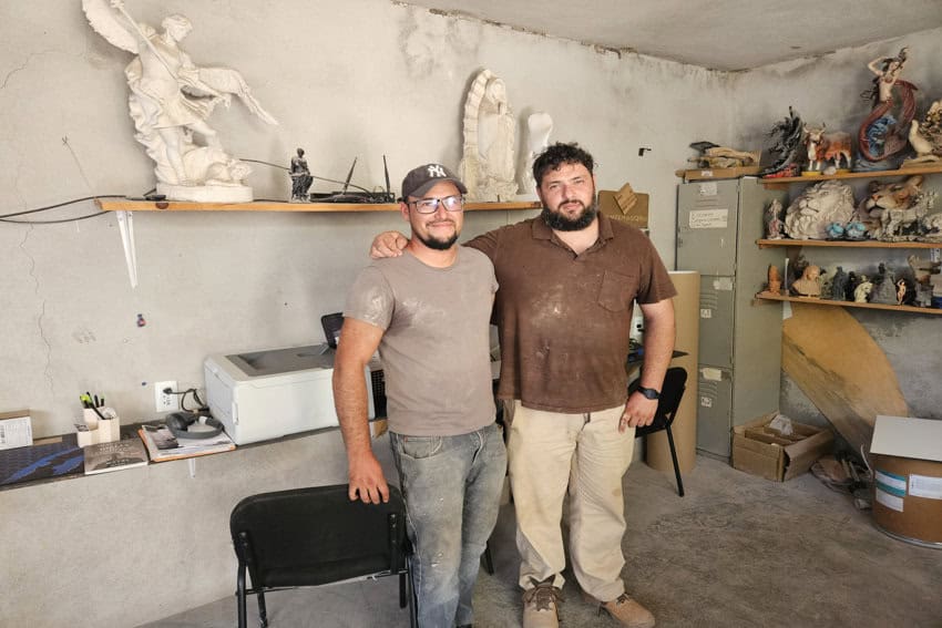 Hugo Uribe (izquierda) y su hermano José Uribe (derecha) de pie juntos en su taller de Canteras Querétaro, con esculturas y materiales en los estantes.