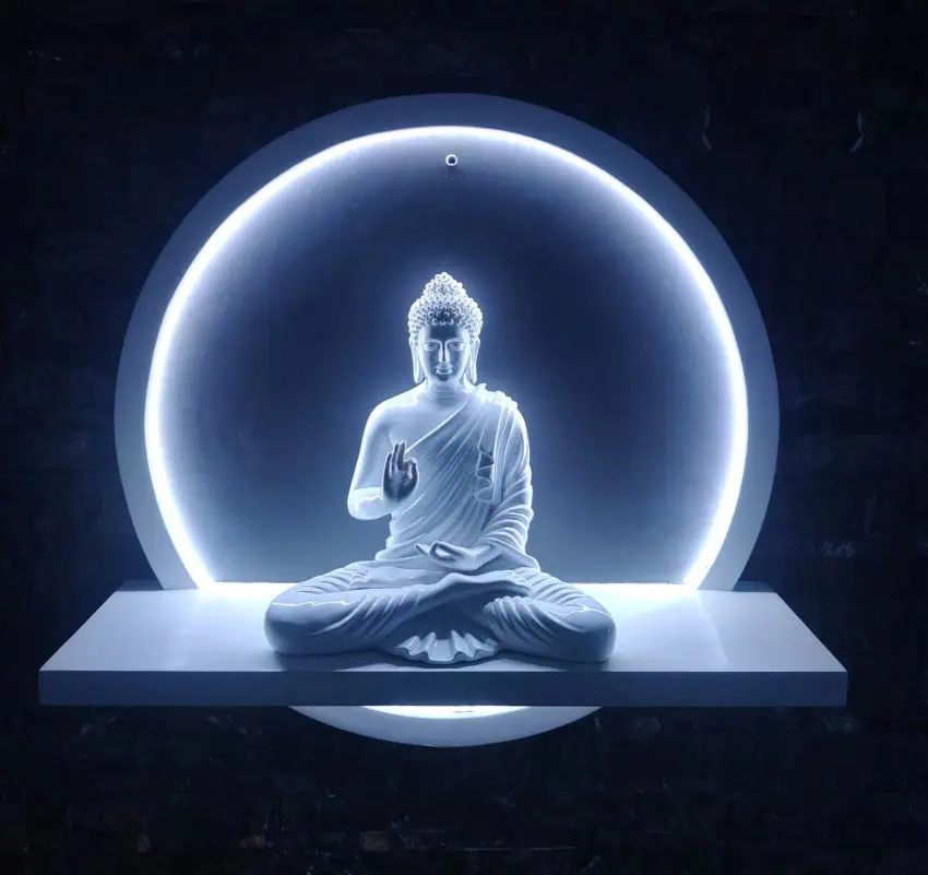 Una estatua blanca de Buda en una pose meditativa, sentada en un estante blanco. Detrás de la estatua, una luz de anillo LED blanca brillante crea un efecto de halo, iluminando la figura contra un fondo oscuro y texturizado.