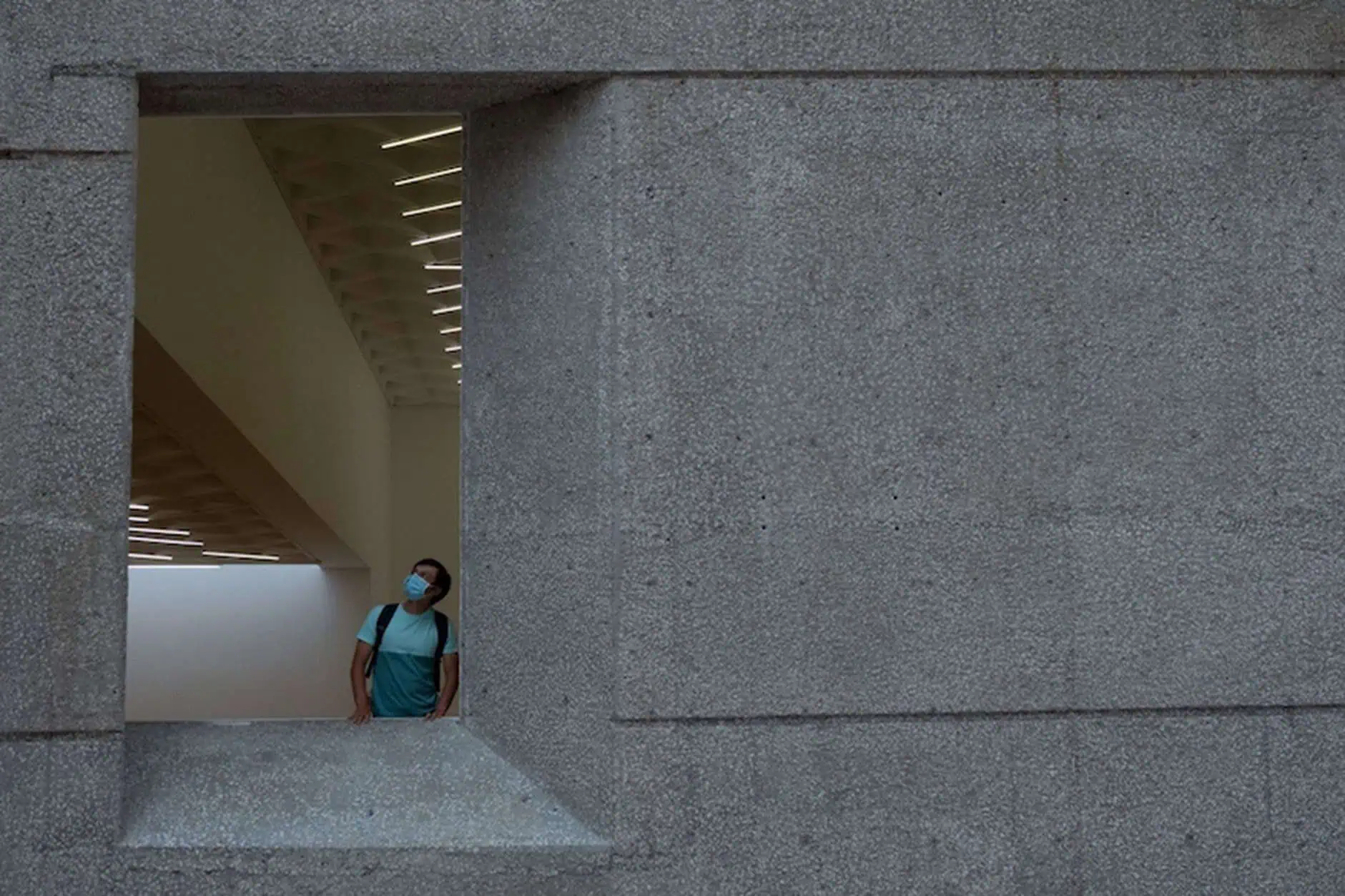 Un hombre que usa una máscara de cara azul claro y una mochila mira hacia arriba y hacia la derecha, enmarcada por una abertura de la ventana de concreto. La ventana es parte de una gran pared de concreto texturizada, y dentro del edificio, se visibles en el techo se visible en el techo.