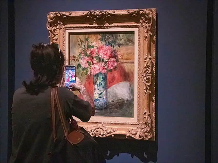 Una persona de espaldas a la cámara toma una foto de una pintura de flores rosadas en un jarrón en una exposición de arte. La pintura está en un marco de oro adornado y representa un ramo de rosas rosadas y otras flores en un jarrón decorativo azul y blanco, ubicado contra un fondo cálido y rojizo.