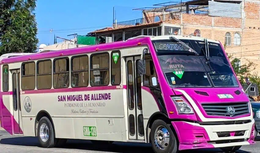 Un autobús de transporte público de color crema y de color magenta está estacionado en una calle en San Miguel de Allende, México. El lado del autobús dice "San Miguel de Allende/Patrimonio de la Humanidad/Cultura, Tradición y Felicidad" y "Ruta 10" es visible en el frente y el lado. En el fondo, los edificios tradicionales mexicanos con techos de azulejos rojos y un poco de follaje verde son visibles debajo de un cielo claro y brillante.