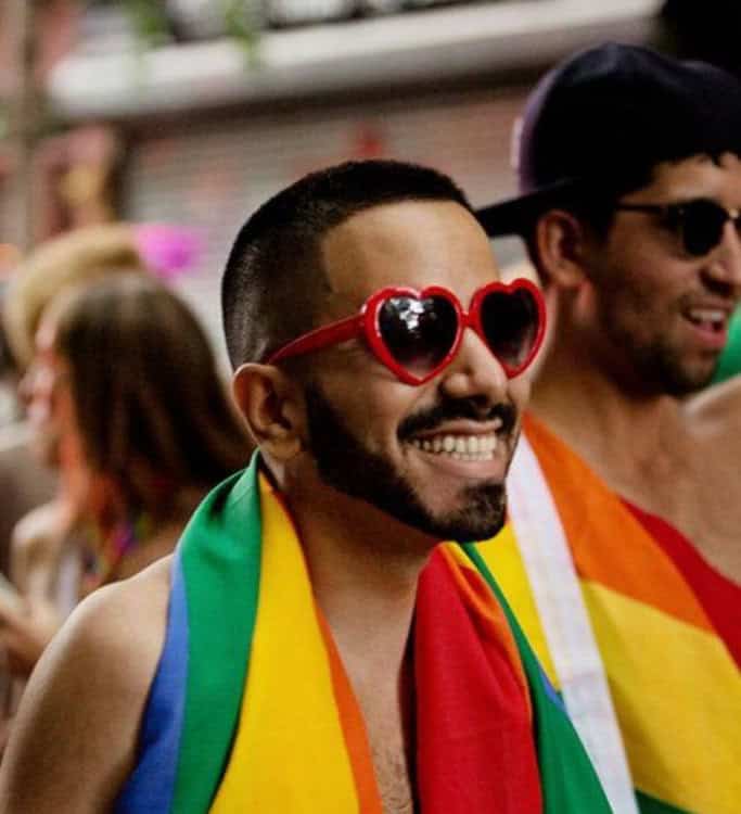 Un tiro en la cabeza de un hombre sonriente con barba y cabello corto, con gafas de sol en forma de corazón rojo y un paño con rayas de arco iris cubiertas sobre sus hombros en un evento de orgullo. Otro hombre con gafas de sol y una bandera del arco iris es parcialmente visible en el fondo de la derecha.