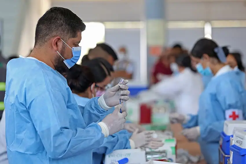 A Mexican man in scrubs and a facemask fills a syringe from a vial in a busy vaccine application center