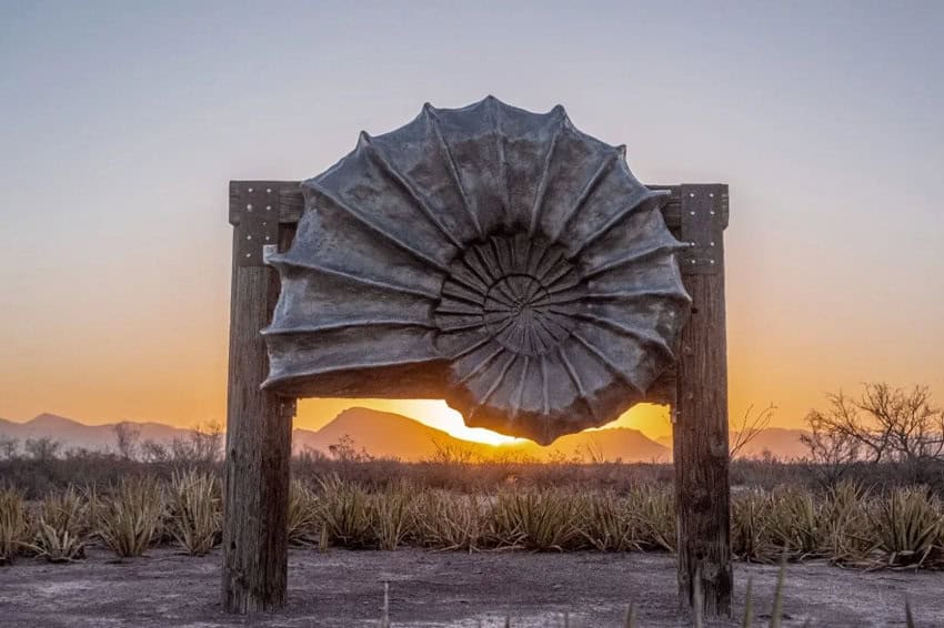 Una gran escultura de metal desgastada que se asemeja a una cáscara de amonita está montada en dos postes de madera, silueta contra una vibrante puesta de sol sobre un paisaje desértico. Las montañas son visibles en la distancia, y la escasa vegetación del desierto llena el primer plano.