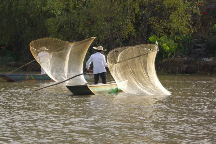 Dos pescadores tradicionales en botes pequeños en un lago tranquilo, cada uno con una red de pesca grande y distintiva de "mariposa" hecha de red de color ligero estirado sobre postes de madera. El pescador en primer plano está en un bote verde y blanco, mirando hacia la izquierda, mientras que el segundo pescador está más atrás en un bote azul, en su mayoría oscurecido. Exuberantes árboles verdes y follaje se alinean en el banco lejano debajo de un cielo brillante.
