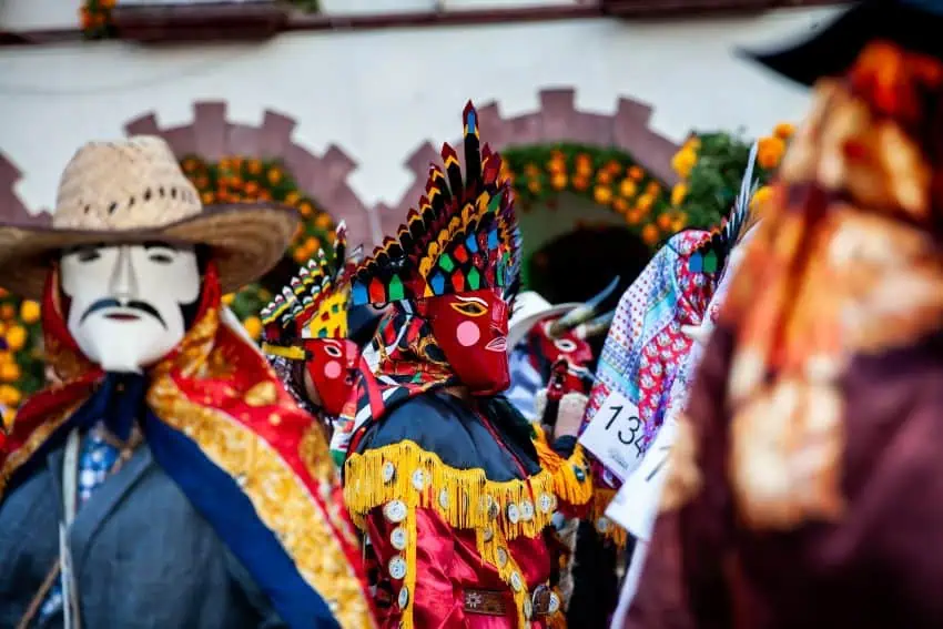 Los artistas y máscaras tradicionales vibrantes, incluida una con una cara roja y tocado emplumado y otro con un sombrero de paja y bigote pintado, participando en una celebración cultural o un desfile en San Luis Potosí, México.