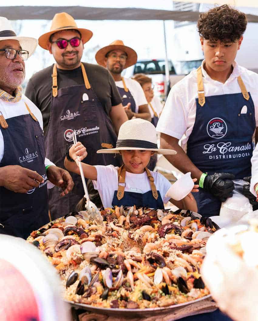 Un gran grupo de chefs y cocineros, algunos con sombreros y delantales, se recogen afuera en una cocina improvisada, de pie alrededor de una enorme paella llena de paella de mariscos. Un niño con un sombrero de paja y un delantal está agitando con entusiasmo la paella, mientras que los adultos observan y ayudan en una concisión en Baja California, México.