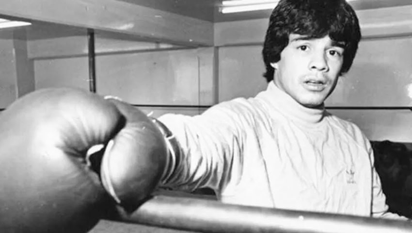 Una foto de primer plano en blanco y negro de un joven boxeador en un anillo de entrenamiento. Tiene el cabello oscuro y ondulado y lleva un cuello de tortuga de manga largo o una camisa de cuello de cuello. Su mano enguantada en boxeo está prominentemente en primer plano, descansando sobre una barra o cuerda, mientras que su mirada se dirige ligeramente fuera de cámara con una expresión enfocada. El fondo parece ser un gimnasio interior con un espejo y accesorios de iluminación flueciente.