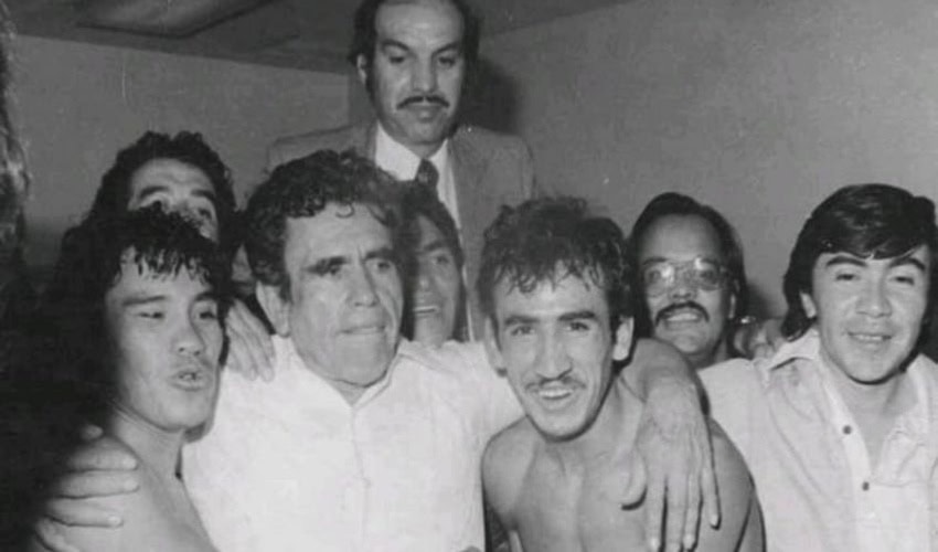 Una fotografía vintage en blanco y negro de un grupo de boxeadores y asociados de boxeo que rodean a Arturo "Cuyo" Hernández, un gerente y promotor mexicano de boxeo. Todos los hombres están reunidos de cerca, muchos con los brazos alrededor del otro. En el centro, un hombre con cabello rizado y una camisa blanca tiene sus brazos alrededor de dos hombres más jóvenes sin camisa con un traje y bigote se mantiene más alto detrás del grupo, mirando sobre sus cabezas.