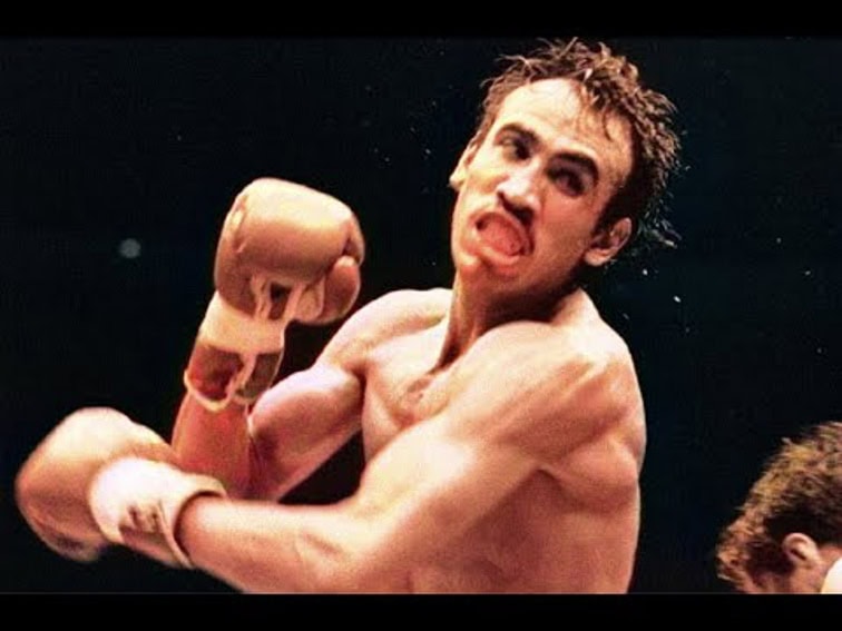 Campeón de boxeo mexicano Carlos Zarate en acción, pelea media. Tiene bigote y cabello corto, oscuro y desordenado. Su boca está abierta, lo que sugiere un esfuerzo o un grito, y sus ojos están intensamente enfocados. Lleva guantes de boxeo de color claro y parece estar lanzando o recibiendo un golpe, con tensión muscular visible en los brazos, los hombros y la cara.