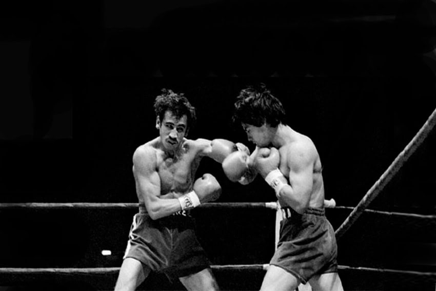 A black-and-white action shot of two boxers exchanging punches in a boxing ring. The boxer on the left, with a mustache and dark, messy hair, is throwing a left hook, while the boxer on the right is in a defensive or counterpunching stance. Both are wearing boxing gloves and shorts.