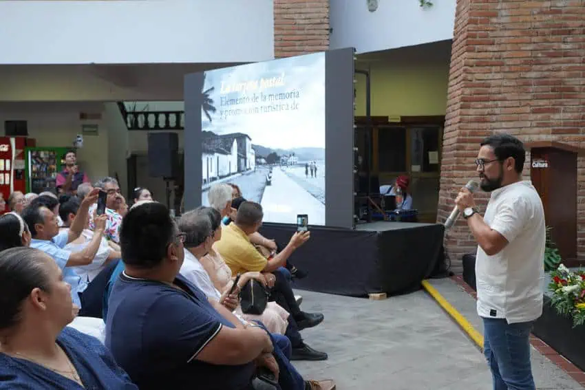 Moisés Hernández López, cronista oficial de Puerto Vallarta, da una conferencia sobre la historia de la ciudad, con una vieja foto de la ciudad proyectada detrás de él y una audiencia escuchando atentamente.