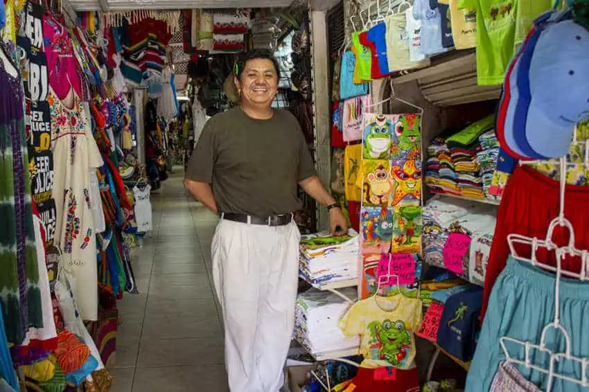 Un proveedor masculino sonriente se encuentra en su estrecho puesto de mercado lleno de coloridas ropa y recuerdos en Puerto Vallarta, México