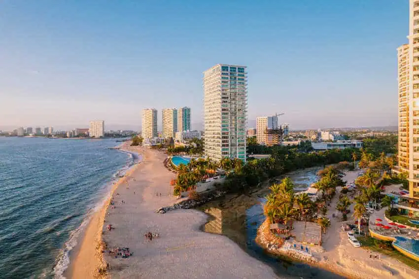 Aerial view of modern high-rise resorts and condominiums lining the sandy beach and coastline of Puerto Vallarta, Mexico, with Banderas Bay stretching into the distance.