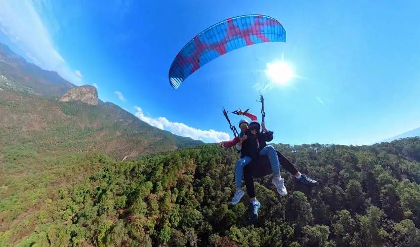 Dos personas en lugar de paraglidismo con un dosel colorido muy por encima de un exuberante bosque verde y montañas bajo un cielo soleado brillante en México