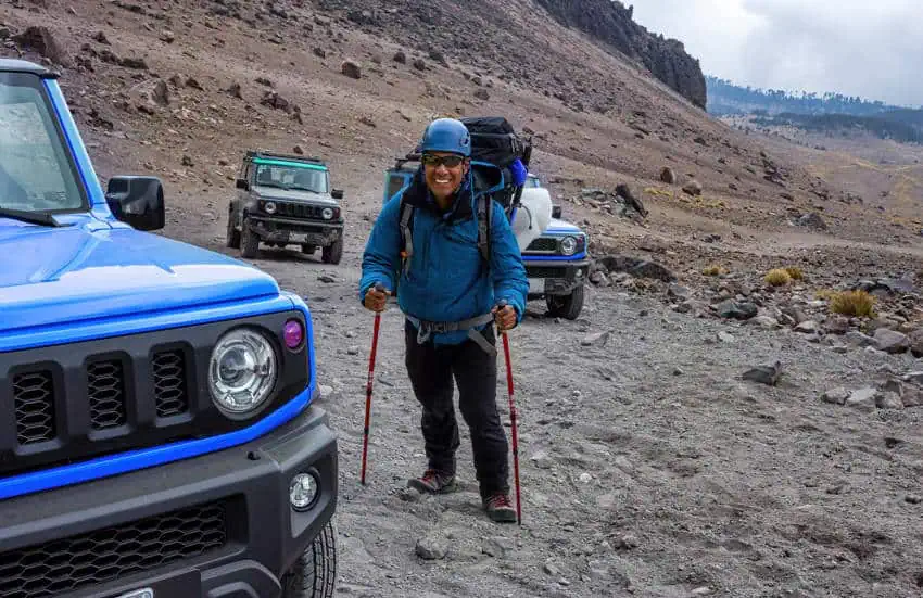 Un montañero sonriente con una mochila y postes de trekking se encuentra en un camino rocoso al lado de un SUV azul en las laderas inferiores de Pico de Orizaba, México