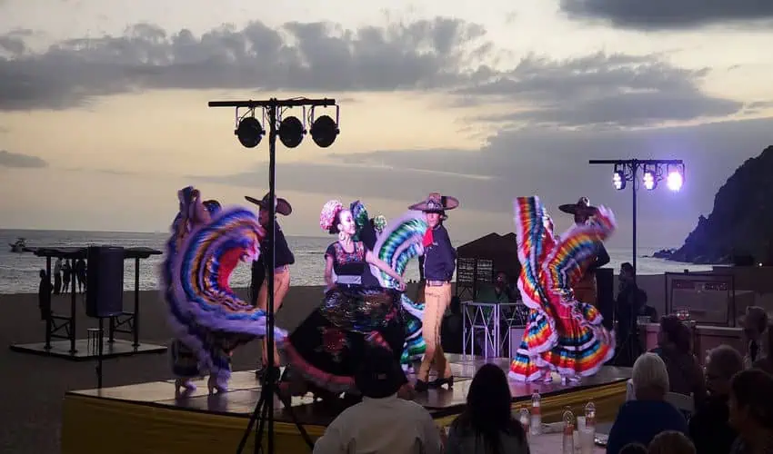 Un grupo de bailarines con coloridos vestidos y sombreros tradicionales mexicanos se presenta en un escenario al aire libre en una playa al anochecer.