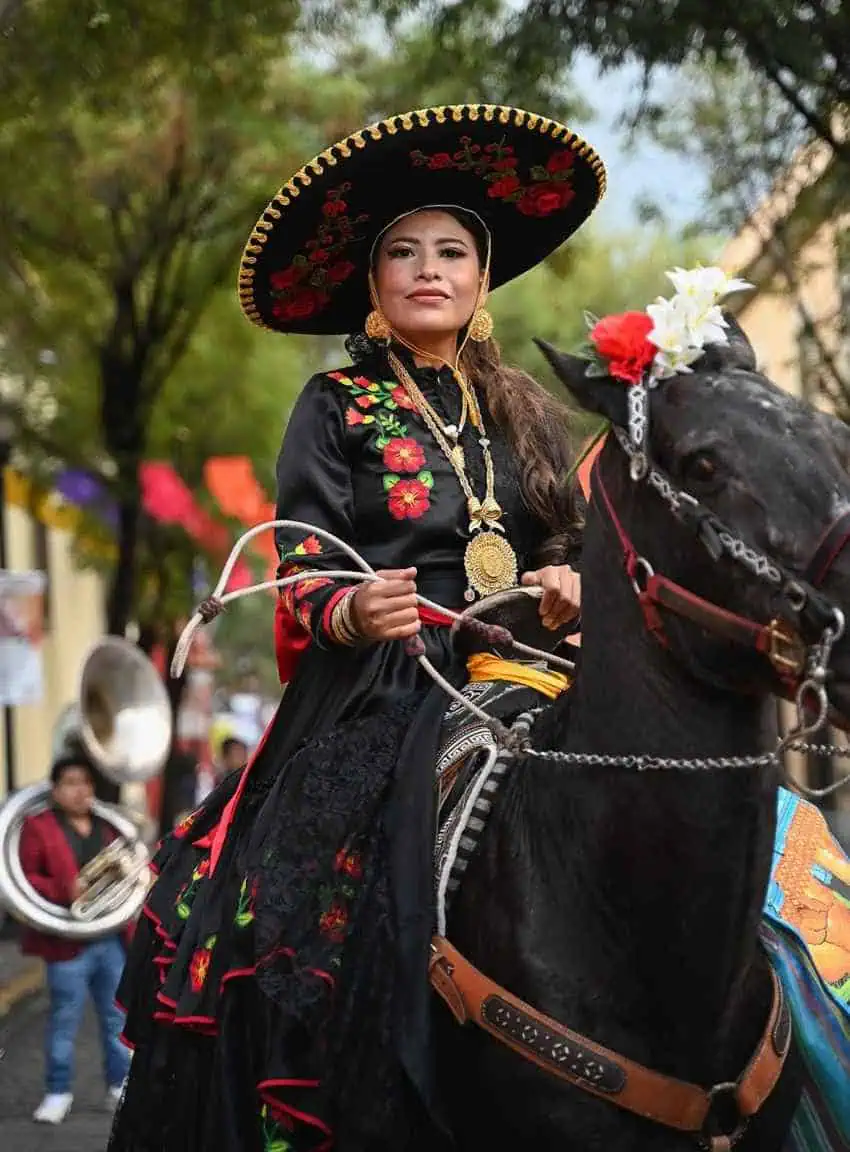 Una mujer con un vestido de charro negro elaboradamente bordado y un sombrero de ala ancha adornada con flores, con grandes joyas doradas, se sienta sobre un caballo oscuro y sostiene riendas, mirando hacia el espectador. Las banderas coloridas se colocan en la calle en el fondo borrosa.