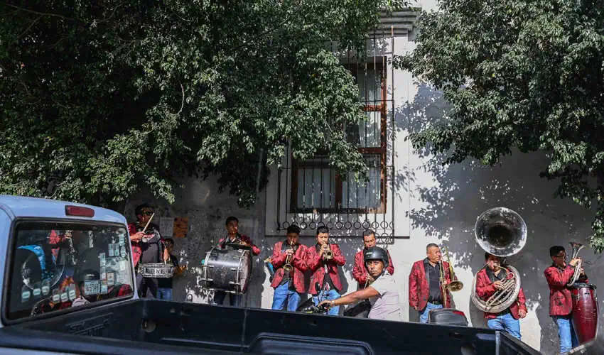 Un grupo de músicos masculinos en chaquetas rojas toca varios instrumentos de latón y tambores en una calle, parcialmente oscurecido por un camión estacionado y árboles verdes, con un edificio blanco en el fondo.