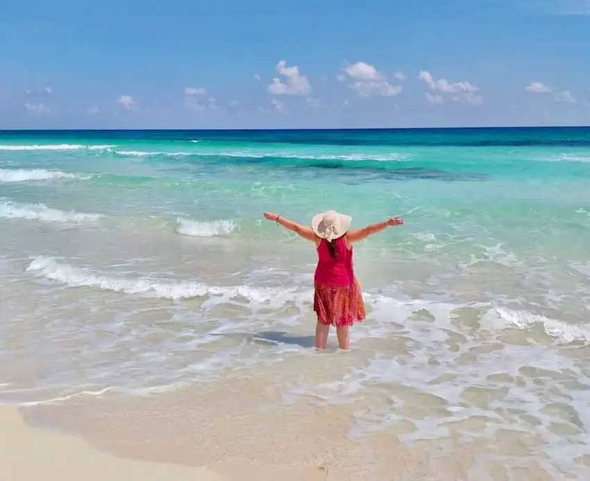 Una mujer con un sombrero de sol y un vestido rojo se encuentra en el agua transparente y turquesa de una playa mexicana, brazos extendidos mirando el horizonte de la vaniduría del océano